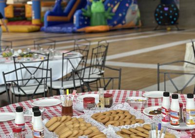Mesa con merienda y dulces en primer plano con varios castillos hinchables de grandes dimensiones instalados al fondo en un pabellón.